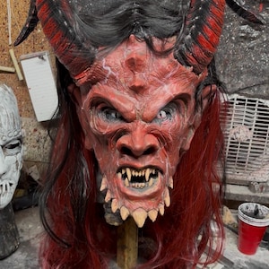 Könnte beinhalten: Eine detaillierte rote Dämonenmaske mit großen, texturierten Hörnern mit schwarzen Akzenten, scharfen weißen Zähnen und einer gefurchten Stirn. Die Maske ist mit langem, glattem schwarzem Haar verziert, das an den Enden in leuchtendes Rot übergeht.