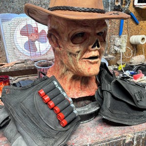 May include: A distressed brown cowboy hat rests on a decaying mask with a skeletal nose. A black tactical vest with red-tipped shotgun shells and a black pouch are in the foreground. The scene evokes a horror or cosplay theme, with a focus on the mask's unsettling details.