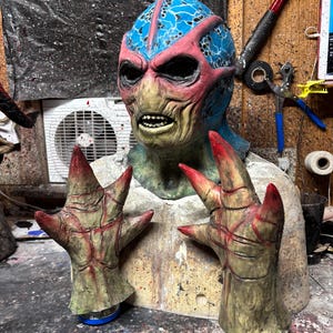 May include: A detailed, handmade monster mask with a blue and pink head, green face, and sharp teeth. The mask is paired with two matching clawed hands with red tips. The mask and hands are mounted on a bust.
