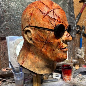 May include: A gruesome, weathered mask with a scarred, orange-brown complexion. The mask features deep cuts and is adorned with black, round sunglasses. The mask is mounted on a wooden stand, suggesting a prop or Halloween decoration.