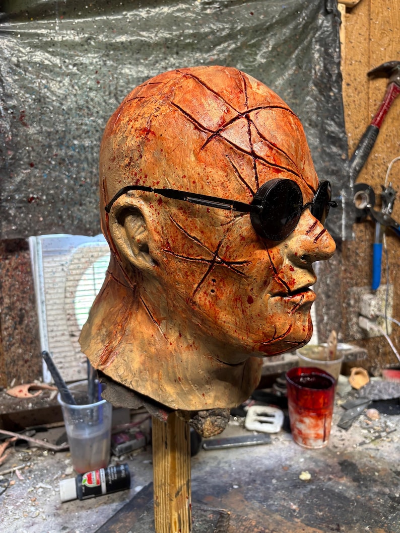 May include: A gruesome, weathered mask with a scarred, orange-brown complexion. The mask features deep cuts and is adorned with black, round sunglasses. The mask is mounted on a wooden stand, suggesting a prop or Halloween decoration.