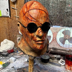 May include: A grotesque, weathered mask with a textured, orange-brown surface, featuring deep, red-stained cuts and black round sunglasses. The mask is mounted on a wooden stand, suggesting a horror or Halloween theme.