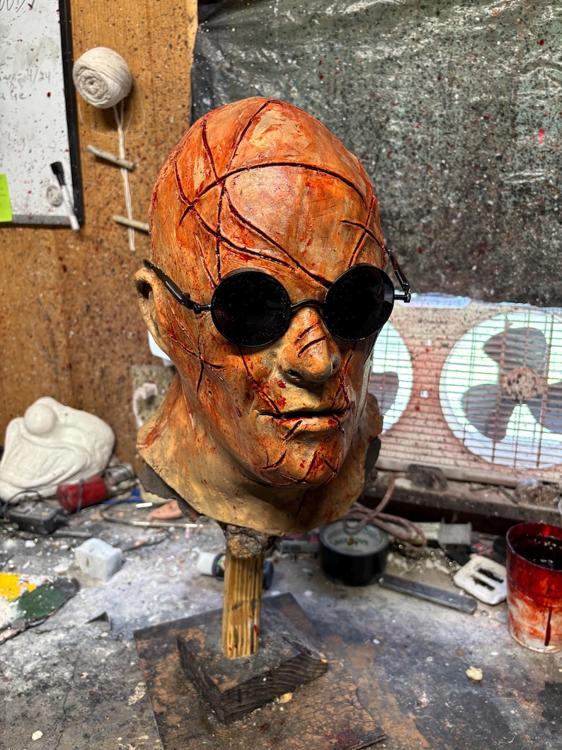 May include: A grotesque, weathered mask with a textured, orange-brown surface, featuring deep, red-stained cuts and black round sunglasses. The mask is mounted on a wooden stand, suggesting a horror or Halloween theme.