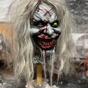 Puede incluir: Una máscara espeluznante con cabello blanco, sangre y una sonrisa, con ojos amarillos. La máscara está en un soporte de madera.