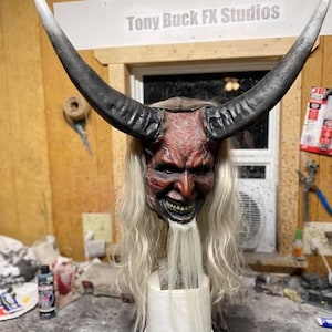 Könnte beinhalten: Eine rote und schwarze Teufelmaske mit langen weißen Haaren und Hörnern. Die Maske befindet sich auf einem weißen Eimer und der Text "Tony Buck FX Studios" ist im Hintergrund sichtbar.