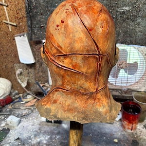 May include: A weathered, orange-brown mask with deep, red-lined cuts across the surface. The mask is mounted on a wooden stand, suggesting a prop or decorative item. The overall aesthetic is reminiscent of horror or Halloween themes.