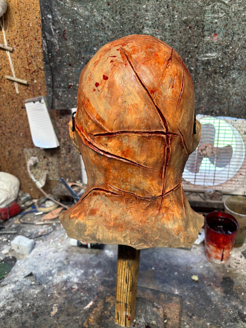 May include: A weathered, orange-brown mask with deep, red-lined cuts across the surface. The mask is mounted on a wooden stand, suggesting a prop or decorative item. The overall aesthetic is reminiscent of horror or Halloween themes.