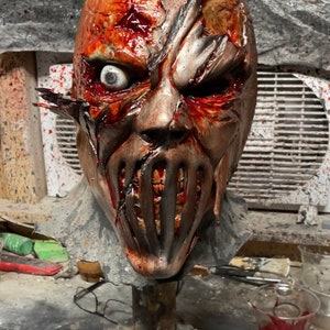 May include: A detailed, realistic horror mask with a torn, bloody face and a metal mouth guard. The mask is mounted on a wooden base and is painted in shades of red, brown, and gray.