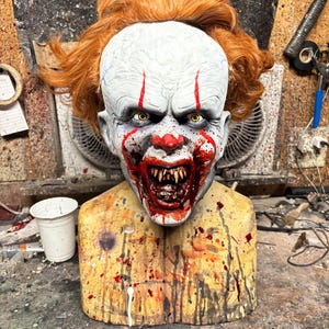 May include: A close-up of a scary clown mask with red and white makeup, a wide open mouth with sharp teeth, and yellow eyes. The mask is on a wooden stand with red and white paint splatters.