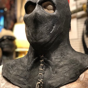 May include: A black leather mask with a stitched neck opening and eye holes. The mask has a textured surface and appears to be made of a thick material.