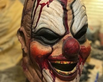 Könnte beinhalten: Eine gruselige Clownsmaske mit einem blutigen, offenen Kopf, der ein Gehirn enthüllt. Die Maske hat ein breites, zahniges Grinsen und eine rote Nase. Die Maske ist aus einem realistischen Material gefertigt und perfekt für Halloween oder einen Horrorfilm.