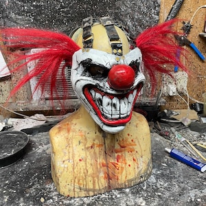 Puede incluir: Una máscara de payaso blanca y roja con una amplia sonrisa de dientes y una nariz roja. La máscara tiene correas negras y cabello rojo. La máscara está en una cabeza de maniquí con una superficie marrón y blanca.