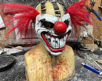Puede incluir: Una máscara de payaso blanca y roja con una amplia sonrisa de dientes y una nariz roja. La máscara tiene correas negras y cabello rojo. La máscara está en una cabeza de maniquí con una superficie marrón y blanca.