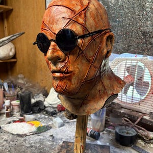 May include: A sculpted head with a textured, orange-brown surface, featuring black round sunglasses. The head is mounted on a wooden stand. The surface has deep cuts and lines. The background includes a sign that says "Famous Hollywood".