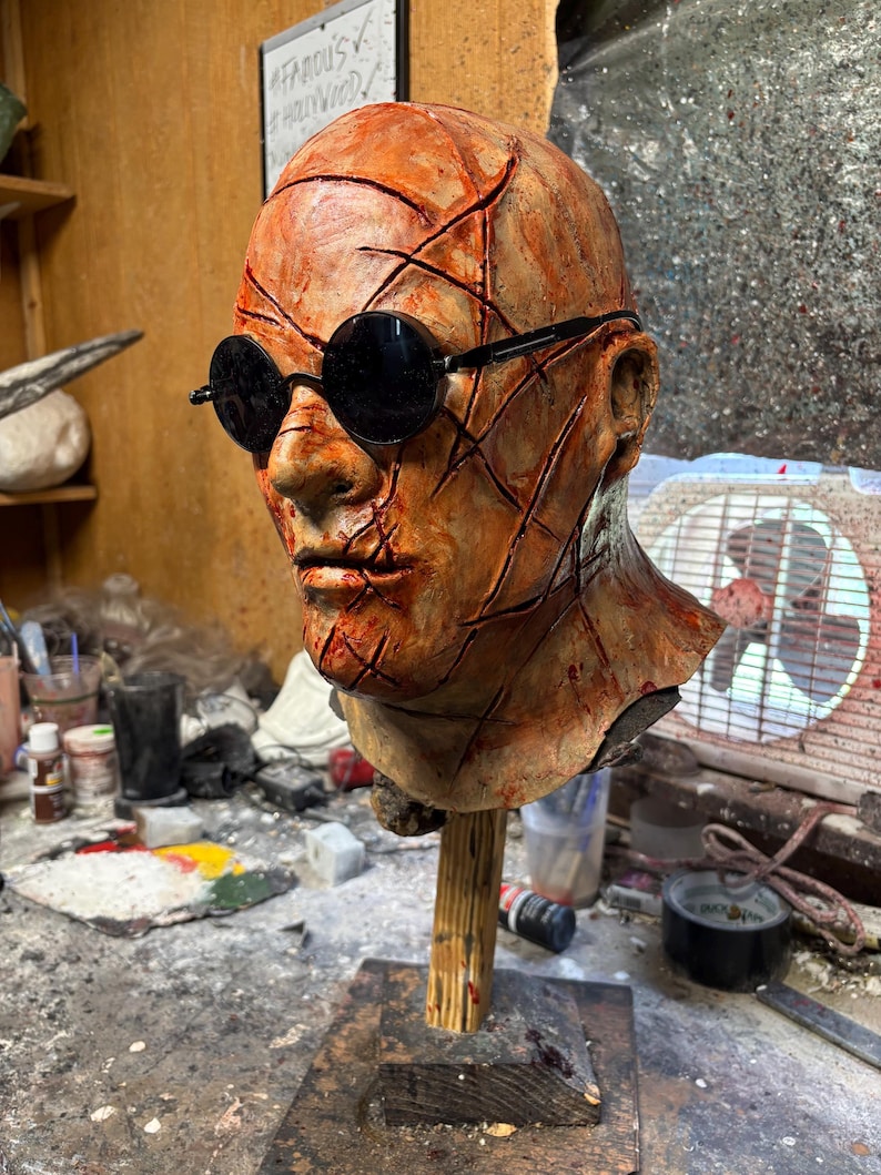 May include: A sculpted head with a textured, orange-brown surface, featuring black round sunglasses. The head is mounted on a wooden stand. The surface has deep cuts and lines. The background includes a sign that says "Famous Hollywood".