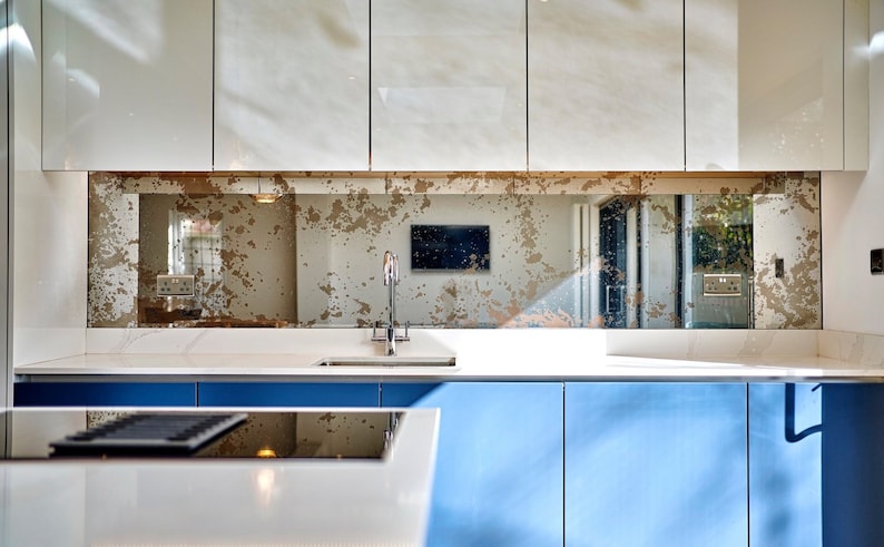 May include: A modern kitchen with white cabinets and a blue countertop. The backsplash is a distressed mirror with a gold finish. A stainless steel faucet is mounted above a white sink.