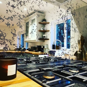 May include: A black gas hob with four burners and a white candle with the word "CALM" on the label. The hob is in a kitchen with a wooden worktop and a window in the background.