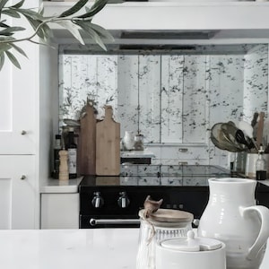 May include: A white kitchen with a black stove and a distressed mirror backsplash. There are various kitchen utensils and a glass jar with a wooden lid on the counter.