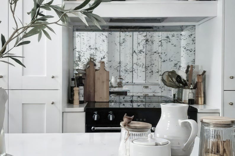 May include: A white kitchen with a black stove and a distressed mirror backsplash. There are various kitchen utensils and a glass jar with a wooden lid on the counter.
