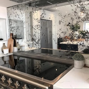 May include: A modern kitchen featuring a stainless steel hob and a backsplash made of antique mirrored tiles. The mirror tiles have a distressed look with a white background and black speckles. The hob is turned off and the surface is clean.