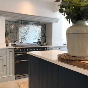 May include: A kitchen with white cabinets, a black range, and a white worktop. The backsplash is a distressed mirror with a gold frame. A vase with green plants sits on the worktop.