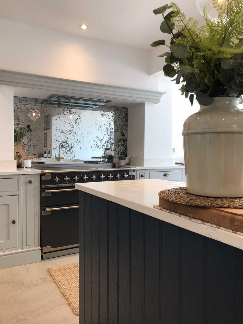May include: A kitchen with white cabinets, a black range, and a white worktop. The backsplash is a distressed mirror with a gold frame. A vase with green plants sits on the worktop.