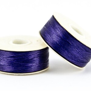 May include: Two spools of dark purple thread are displayed against a white background. The thread is tightly wound around a white cardboard spool. The spools are slightly angled, showcasing the thread's texture and color.