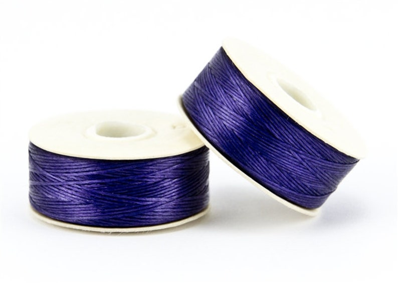May include: Two spools of dark purple thread are displayed against a white background. The thread is tightly wound around a white cardboard spool. The spools are slightly angled, showcasing the thread's texture and color.