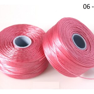 Pode incluir: Dois carret&eacute;is de fio rosa, cada um enrolado em torno de uma bobina de pl&aacute;stico branca. Os carret&eacute;is s&atilde;o etiquetados com "06 - Rosa".
