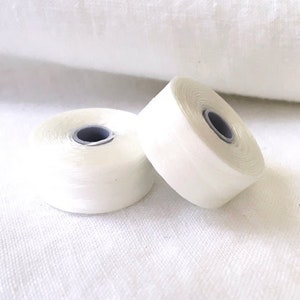 May include: Two spools of white thread, each with a gray core, rest on a white fabric surface. The thread is tightly wound, and the spools are positioned at slightly different angles, showcasing their cylindrical shape.