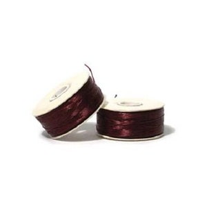 May include: Two spools of burgundy-colored thread are displayed against a white background. The thread is tightly wound around white cardboard spools. The image is well-lit, highlighting the texture and color of the thread.