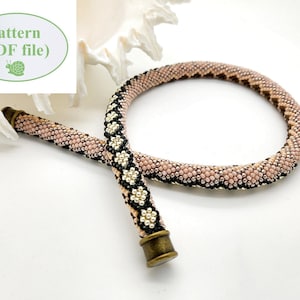 May include: A beaded necklace with a pink, black, and silver pattern. The necklace is made of small beads and has a metal clasp. The necklace is lying on a white surface.