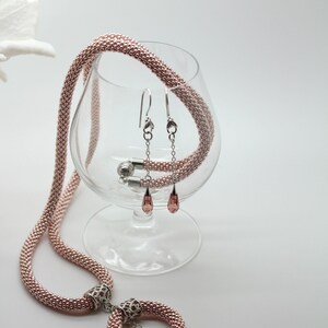 May include: A rose gold beaded necklace with a circular pendant and a teardrop-shaped crystal. Matching rose gold beaded earrings with teardrop-shaped crystals are also included. The jewelry is displayed on a glass pedestal.