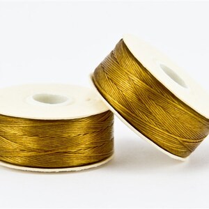 May include: Two spools of golden-colored thread on white spools. The thread is tightly wound, and the spools are set against a white background. The thread has a slight sheen, suggesting a satin or silk material, ideal for crafts.
