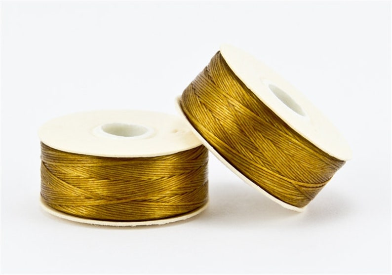 May include: Two spools of golden-colored thread on white spools. The thread is tightly wound, and the spools are set against a white background. The thread has a slight sheen, suggesting a satin or silk material, ideal for crafts.