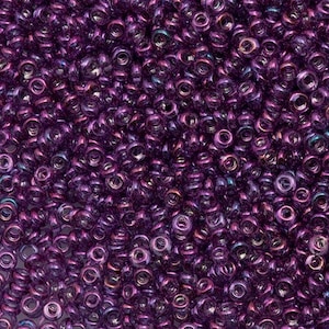 Puede incluir: Primer plano de un montón de pequeñas cuentas de semillas de color púrpura oscuro. Las cuentas tienen un brillo metálico, con toques de colores iridiscentes como el azul y el dorado. Ideales para la fabricación de joyas y proyectos de artesanía.