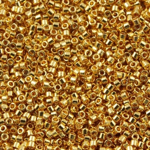 May include: A close-up image of a large quantity of gold-coloured seed beads. The beads are small and round, with a hole in the centre. They are arranged in a random pattern, creating a textured surface.