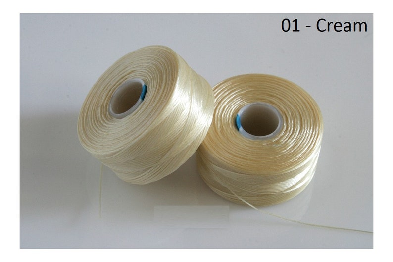 Pode incluir: Dois carret&eacute;is de fio de cor creme. O fio &eacute; enrolado em torno de um carretel de pl&aacute;stico branco com um centro azul.