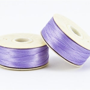 May include: Two spools of lavender-colored thread. The thread is tightly wound around a beige cardboard core. One spool is upright, while the other is tilted, showing the thread's texture. Ideal for crafting and sewing projects.
