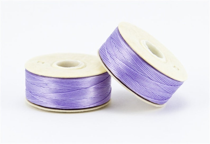 May include: Two spools of lavender-colored thread. The thread is tightly wound around a beige cardboard core. One spool is upright, while the other is tilted, showing the thread's texture. Ideal for crafting and sewing projects.
