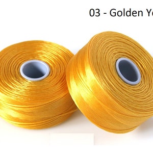 Pode incluir: Dois carret&eacute;is de fio amarelo dourado, cada um enrolado em torno de um carretel central de pl&aacute;stico branco.