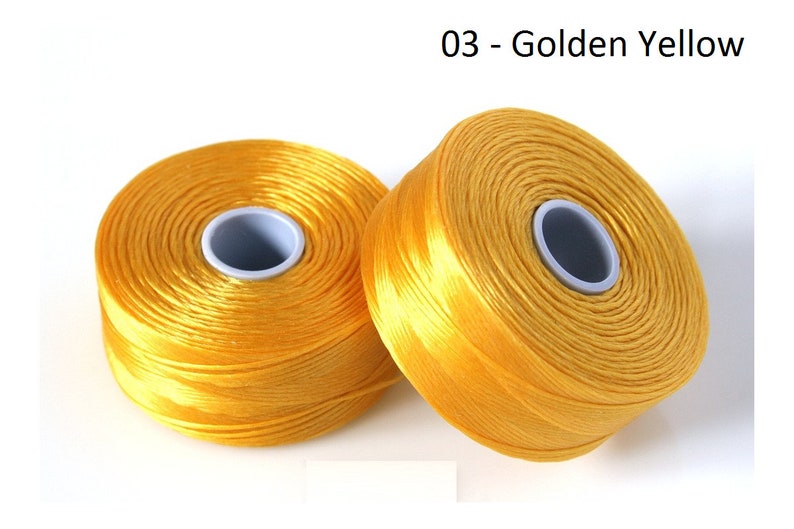 Pode incluir: Dois carret&eacute;is de fio amarelo dourado, cada um enrolado em torno de um carretel central de pl&aacute;stico branco.