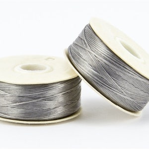 May include: Two spools of gray thread are displayed against a white background. The thread is tightly wound around a cream-colored cardboard spool. One spool is upright, while the other is tilted, creating a dynamic composition.