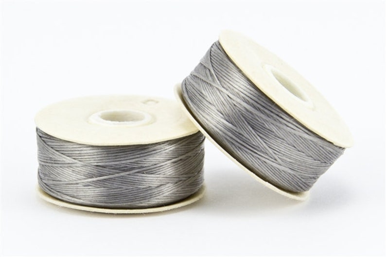 May include: Two spools of gray thread are displayed against a white background. The thread is tightly wound around a cream-colored cardboard spool. One spool is upright, while the other is tilted, creating a dynamic composition.