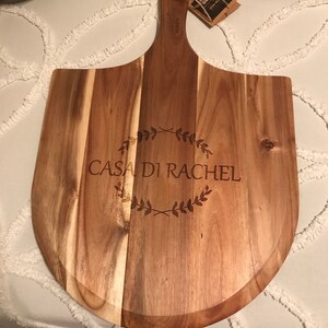 May include: A wooden pizza peel with a handle, engraved with the words "Casa di Rachel" and a wreath design. The peel is made of acacia wood.