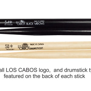 May include: Two pairs of drumsticks, one black and one natural wood. The black pair is labeled "5A Red Hickory" and "Los Cabos Black Widows". The natural wood pair is labeled "5B Hickory" and "Los Cabos Drumsticks". Both pairs have a small Los Cabos logo on the back of each stick.