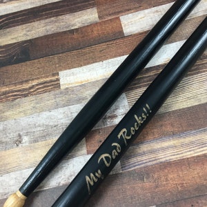 May include: A pair of black wooden drumsticks with a gold inscription that reads "My Dad Rocks!"