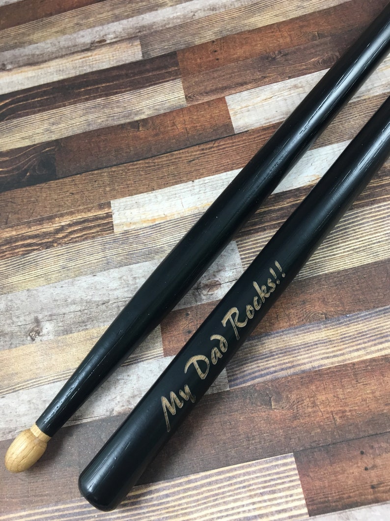 May include: A pair of black wooden drumsticks with a gold inscription that reads "My Dad Rocks!"