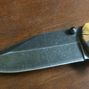 Puede incluir: Una navaja de bolsillo plegable con una hoja negra y un mango de camuflaje. La hoja est&aacute; parcialmente abierta, revelando su filo. El mango presenta un patr&oacute;n de marr&oacute;n, verde y beige, y la navaja est&aacute; sobre una superficie de madera.