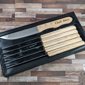 May include: A black box with a set of 6 steak knives with wooden handles. The knife on top has the name "Chef Alex" engraved on the handle.
